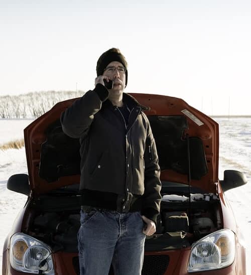 A man calling for help on the side of a snowy road with the hood of his vehicle open
