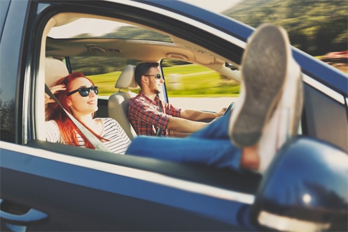 7 Things to Do During 2023 Spring Car Care Month in Amagansette, NY. Image of young couple taking a drive with windows open on a nice spring day. They are wearing sunglasses and the passenger has her feet propped up on side view mirror.